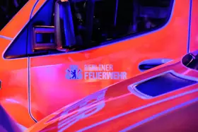 Nahaufnahme der Fahrertür eines orange-roten Fahrzeugs mit dem Schriftzug 'Berliner Feuerwehr' und einem Wappen.