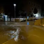 Überschwemmte Baustelle bei Nacht mit Bauzaun, mobiler Toilette, Straßenlaterne und einem Haufen Erde vor einem mehrstöckigen Gebäude.