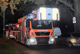 Zwei rot-orange Feuerwehrfahrzeuge mit Blaulicht stehen nachts auf einer Straße, rechts geparkte Autos und ein Halteverbotsschild.