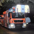 Zwei rot-orange Feuerwehrfahrzeuge mit Blaulicht stehen nachts auf einer Straße, rechts geparkte Autos und ein Halteverbotsschild.