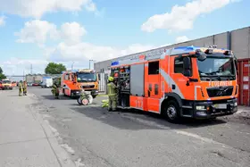 Zwei orange-rote Feuerwehrfahrzeuge stehen auf einem Industriehof, Feuerwehrleute in Schutzkleidung arbeiten daneben.