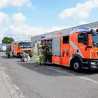 Zwei orange-rote Feuerwehrfahrzeuge stehen auf einem Industriehof, Feuerwehrleute in Schutzkleidung arbeiten daneben.