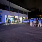 Mehrere Personen in Schutzkleidung stehen vor einem beleuchteten ALDI-Eingang bei Nacht, daneben ein geparktes Auto und ein Fahrrad.