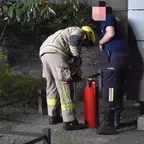 Zwei Männer, einer in Feuerwehrschutzkleidung mit Helm, der andere in dunkler Arbeitskleidung, bedienen gemeinsam eine rote Kübelspritze vor einem Gebäude.