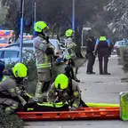 Mehrere Feuerwehrleute mit gelben Helmen und Atemschutzgeräten bereiten eine Trage auf einem Gehweg vor, im Hintergrund stehen Polizisten und ein Rettungswagen mit Blaulicht.