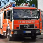 Orangerotes Feuerwehrfahrzeug der Marke MAN mit blauen Rundumleuchten, geparkt vor einem Gebäude.