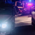 Silberner Pkw mit geöffneter Fahrertür steht nachts vor einem Polizeiauto mit eingeschaltetem Blaulicht und Anhaltesignalgeber, daneben steht eine Person.