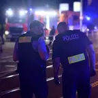 Zwei Polizisten in dunkler Kleidung mit gelben "POLIZEI"-Schriftzügen stehen auf einer Straße bei Nacht, im Hintergrund Blaulicht und Einsatzfahrzeuge.