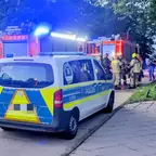 Polizeiwagen mit eingeschaltetem Blaulicht steht vor zwei Feuerwehrfahrzeugen, mehrere Feuerwehrleute in Schutzkleidung stehen auf der Straße.