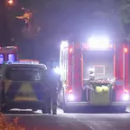 Ein Einsatzfahrzeug der Feuerwehr mit eingeschalteten Lichtern steht auf einer dunklen Straße, daneben ein Polizeiauto und eine stehende Person.