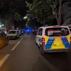 Polizeifahrzeuge mit Blaulicht stehen nachts auf einer Straße, mehrere Personen sind im Hintergrund sichtbar.