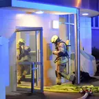 Drei Feuerwehrleute in Schutzkleidung und Helmen vor einem Gebäude mit offenem Glastürrahmen bei Nacht. Davor liegen Schläuche auf dem Boden.