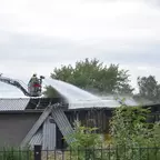 Feuerwehrmann auf einer Drehleiter spritzt Wasser auf ein Flachdach eines Gebäudes, umgeben von Bäumen und Sträuchern.