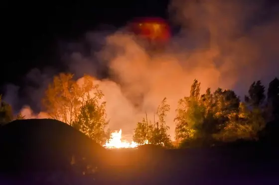 Feuer hinter Bäumen in der Nacht, Rauch steigt auf, dunkle Silhouetten im Vordergrund.