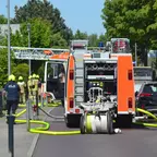 Feuerwehrauto mit ausgefahrener Leiter und mehreren Feuerwehrleuten in Schutzkleidung auf einer Straße in Wohngebiet.