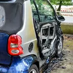 Kleines Auto mit geschmolzener und stark beschädigter Beifahrertür, Rücklicht und Heckpartie intakt, steht auf nassem Boden neben einer Straße.