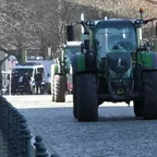 Traktor fährt auf gepflasterter Straße in einer städtischen Umgebung, im Hintergrund weitere Fahrzeuge und Bäume.