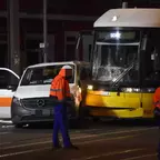 Straßenbahn mit zerbrochenem Frontfenster steht neben einem weißen Mercedes-Van mit geöffneter Fahrertür, zwei Personen in orange-blauer Arbeitskleidung stehen auf der Straße.