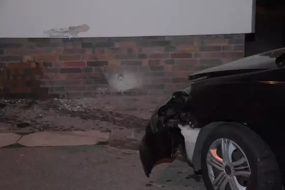 Vorderer Bereich eines schwarzen Fahrzeugs mit beschädigtem Kotflügel vor einer Hauswand mit sichtbarer Einschlagstelle.