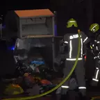 Zwei Feuerwehrleute in Schutzkleidung und Helm stehen nachts vor einem geöffneten Kleidercontainer, aus dem Müllsäcke herausragen.