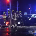 Mehrere Feuerwehrleute in Schutzkleidung stehen nachts an einer Unfallstelle mit einem beschädigten Auto und Einsatzfahrzeugen mit Blaulicht.