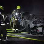 Zwei Feuerwehrleute mit gelben Helmen löschen nachts einen ausgebrannten Wagen mit einem Schlauch.