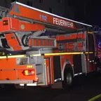 Feuerwehr-Drehleiterfahrzeug bei Nacht, im Hintergrund Feuerwehrleute und Blaulicht.