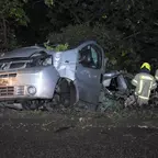 Zerstörter silberner Kastenwage steht nachts an einem Baum, zwei Feuerwehrleute in Schutzkleidung arbeiten am Fahrzeug.