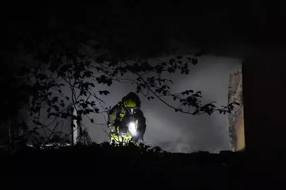 Feuerwehrmann in Schutzkleidung mit Helm und Taschenlampe in dunkler, nebliger Umgebung, umgeben von Pflanzen und einer Mauer.