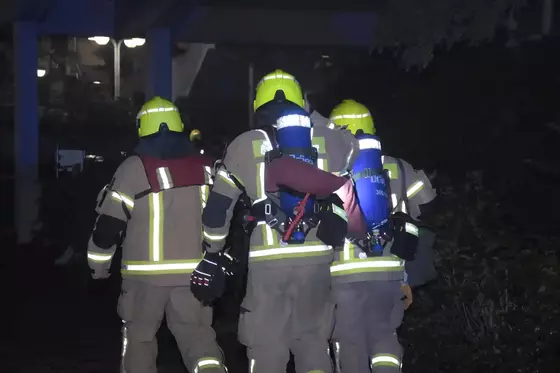 Drei Feuerwehrleute mit gelben Helmen und zwei davon mit Atemschutzgeräten, gehen bei Dunkelheit, weg vom Betrachter.