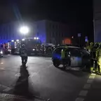 Nächtliche Unfallstelle mit mehreren Feuerwehrleuten in Schutzkleidung neben zwei beschädigten Autos an einer Kreuzung, Einsatzfahrzeuge mit Blaulicht im Hintergrund.