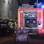 Feuerwehrauto mit offenem Geräteraum und Blaulicht steht in einer dunklen Straße, mehrere Feuerwehrleute in Schutzkleidung stehen daneben.
