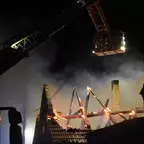 Brennendes Dach eines Gebäudes bei Nacht, Drehleiter mit ausgefahrenem Korb im Einsatz, Rauch steigt auf.