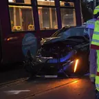 Unfall mit einem schwarzen Auto, das frontal gegen eine Straßenbahn geprallt ist, mehrere Einsatzkräfte in Warnkleidung stehen daneben.