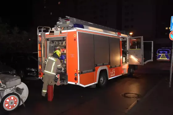 Feuerwehrmann in Schutzkleidung an einem roten Feuerwehrfahrzeug mit geöffneten Fächern bei Nacht. Eine Kübelspritze steht hinter dem Fahrzeug.