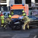 Feuerwehrleute schieben ein schwarzes Auto nach einem Unfall an, im Hintergrund ein Rettungswagen und weitere Einsatzkräfte.