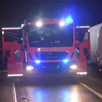 Rotes Feuerwehrfahrzeug mit eingeschalteten Blaulichtern und geöffneten Türen steht nachts auf der Straße zwischen geparkten Fahrzeugen.