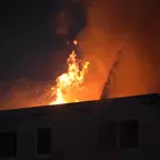 Flammen schlagen aus dem Dach eines Gebäudes in der Nacht, Rauch steigt auf.