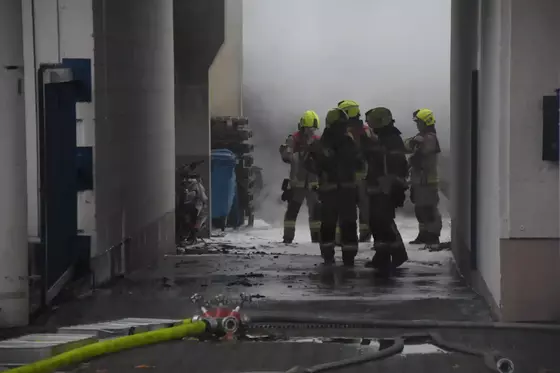 Mehrere Feuerwehrleute mit gelben Helmen stehen in einem schmalen, verrauchten Durchgang zwischen Gebäuden.