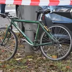 Grünes Fahrrad lehnt an Laternenmast, im Hintergrund parkendes Auto und Absperrband.