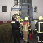Mehrere Feuerwehrleute in Schutzkleidung und Helmen stehen vor einem Wohnhaus mit einem außen angebrachten Aufzugsschacht.