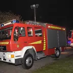 Zwei Feuerwehrfahrzeuge stehen nachts auf einem gepflasterten Weg, ein Feuerwehrmann in Schutzkleidung nähert sich dem hinteren Fahrzeug.