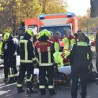 Mehrere Feuerwehrleute und eine Polizistin stehen um eine liegende Person hinter einer Trage vor einem Rettungswagen und einem Bus auf einer Straße.
