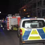 Polizeifahrzeug und Feuerwehrfahrzeuge mit Blaulicht auf einer Straße neben einem beleuchteten Baugerüst bei Nacht.
