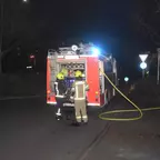 Zwei Feuerwehrleute in Schutzkleidung stehen nachts vor einem Einsatzfahrzeug mit eingeschaltetem Blaulicht und ausgerolltem Schlauch auf einer Straße.