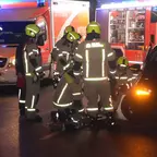 Mehrere Feuerwehrleute in Schutzkleidung und Helmen arbeiten an einem Einsatz neben einem schwarzen Auto und einem Rettungswagen.
