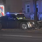 Schwarzer PKW steht gegen ein Absperrgitter an einer Straße, im Hintergrund Rettungswagen und zwei Einsatzkräfte in Schutzkleidung.