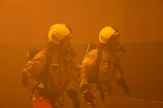 Zwei Feuerwehrmänner in Schutzanzügen und Atemschutzgeräten bewegen sich durch einen verrauchten Tunnel.