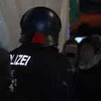 Polizist mit Helm und reflektierender Aufschrift 'POLIZEI' auf dem Rücken steht inmitten einer Menschenmenge am Abend.