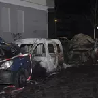 Zwei ausgebrannte Fahrzeuge stehen nachts an einer Hauswand, beide mit geöffneten Motorraum, ebenso beide stark beschädigt.